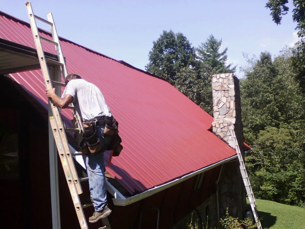 Roofer on a ladder beside a red metal roof and stone chimney — Soffit & Fascia Repair by Roofing Brothers in Riverbank